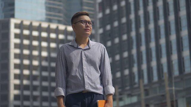 Portrait handsome asian businessman holding a book standing looking around urban city. Slow motion low angle shoot.