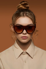 Cropped front view shot of lady, wearing shirt. The girl with bun and wavy hair locks in wrap sunglasses with dark brown rim and gradient lenses. The woman is looking at camera over brown background.