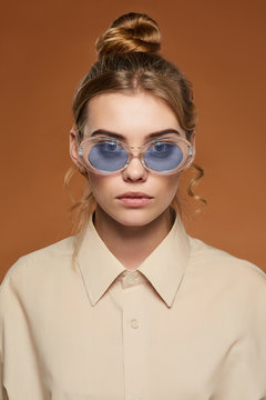 Cropped front view shot of lady with fair hair, wearing shirt. The girl with bun and wavy hair locks in oval-shaped glasses with glitter in blue lenses, is looking at camera on the brown background.