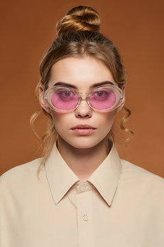 Cropped front view shot of lady with fair hair, wearing shirt. The girl with bun and wavy hair locks in oval-shaped glasses with glitter in pink lenses, is looking at camera on the brown background.