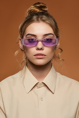 Cropped front view shot of blonde lady, wearing shirt. The girl with bun and wavy hair locks in light purple rectangle sunglasses toreutics on rim, is looking at camera against the sandy background.