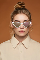 Cropped front view shot of lady, wearing shirt. The girl with bun and wavy hair locks in cat eye-shaped sunglasses with white rim and chameleon lenses, is looking at the camera on brown background.