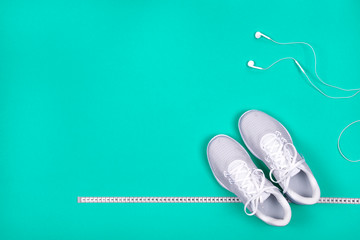 Top view of white sport shoes (sneakers) with measuring tape and white earphones (headphones) on blue-green background.