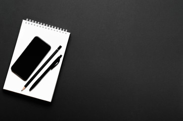 White open notebook with black pencil, pen and smartphone on black background. Office desk, stationery. Top view, flat lay, copy space.