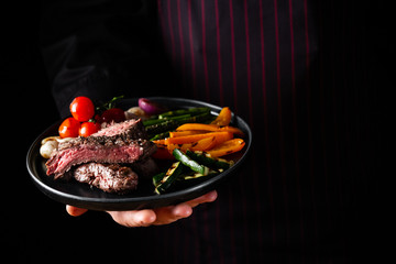 Grilled and sliced beef steak with grilled vegetables served on black plate on black background presentation in chef hands