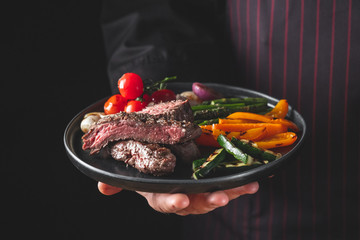 Grilled and sliced beef steak with grilled vegetables served on black plate on black background presentation in chef hands