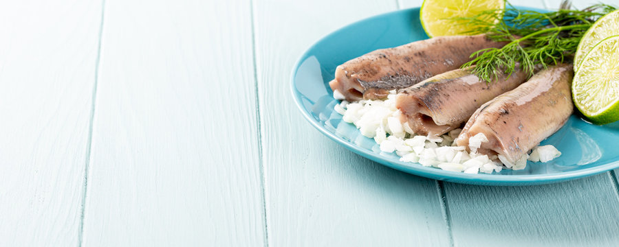 Traditional dutch food freshly salted herring fish with onion called hollandse nieuwe on turquoise plate and wooden background. European food concept with copy space. Banner.