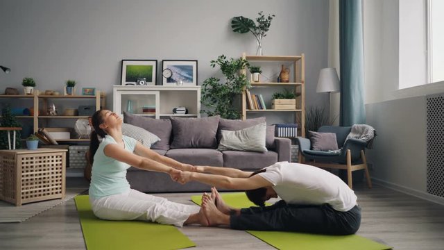 Man and woman young married people are doing stretching exercises holding hands sitting on mats at home relaxing. Active lifestyle and healthcare concept.