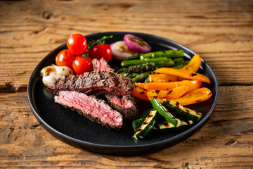 Grilled and sliced beef steak with grilled vegetables served on black plate on old rustic wood background.