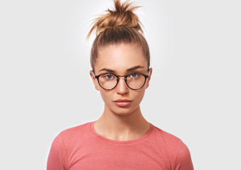 Obraz premium Portrait of attractive blonde young woman wearing round transparent spectacles and casual pink blouse, looks seriously directly into camera, poses against white studio background. People and emotions