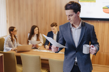 A stylish man in a jacket and a shirt with a cup of coffee in his hand stands and reads documents on the background of working colleagues at office