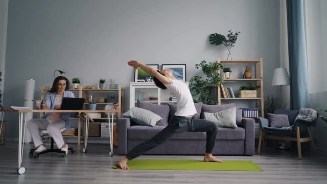 Wife attractive young woman is working with laptop at table while sporty husband is exercising doing yoga on floor in room. People, apartment and hobby concept.