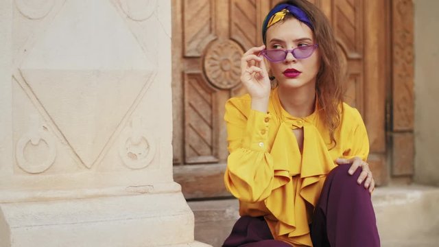 Outdoor fashion portrait of young beautiful elegant lady wearing purple sunglasses, headband, yellow blouse, posing in street of european city