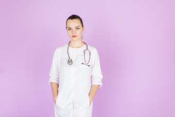 Young female blonde doctor with her hands in pockets with stethoscope on her neck on pink background with space for text. Doctor looks at camera