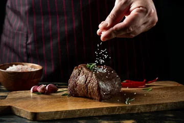 Selbstklebende Fototapeten Chef hands cooking meat steak and adding salt and pepper on black copy space background for menu restaurant or recipe text. © Anton
