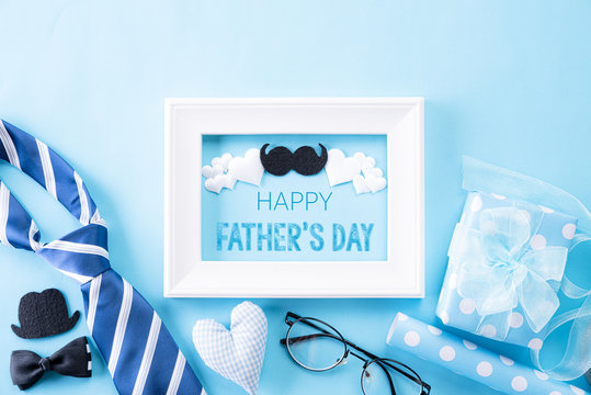 Happy fathers day concept. Top view of blue tie, beautiful gift box, white picture frame with Happy father's day text on bright blue pastel background. Flat lay.