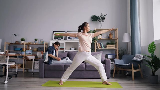 Pretty girl is doing yoga asanas whileyoung man husband is working with laptop talking on mobile phone in nice cozy apartment. Modern lifestyle and sports concept.