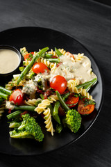 Salad with pasta fusilli, sauce and vegetables in black plate background.