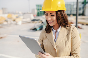 Attractive Caucasian female architect dressed smart casual and with helmet on head using tablet while standing on construction site.