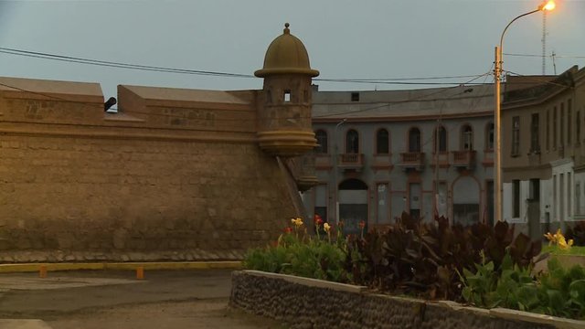 Medium, outer view panning shot of Real Felipe Fortress showing old canon guns in wall positions, high defence wall, beautiful flower garden, street-light, and adjacent buildings, Callao, Lima, Peru.