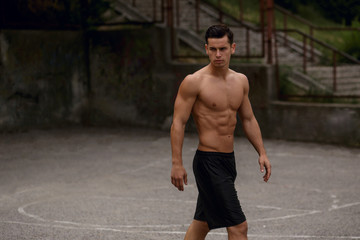Handsome shirtless young sportive man, sitting on bascketball court, showing his six pack abs, toned body, in day time.