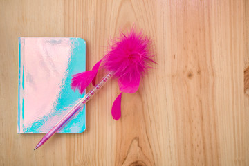 Fototapeta na wymiar Pretty notepad in bright blue, with pink pen that has feathers, isolated on wooden background.