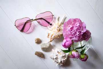 Pink glasses, peonies and sea shells on a white wooden table. Romantic summer flat lay.