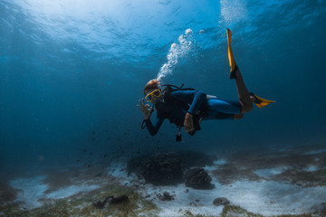 Woman scuba diver swims underwater over the sea bottom exhales the air and shows the OK diving sign