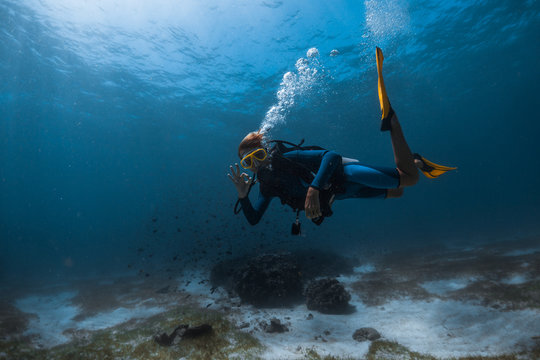 Woman Scuba Diver Swims Underwater Over The Sea Bottom Exhales The Air And Shows The OK Diving Sign