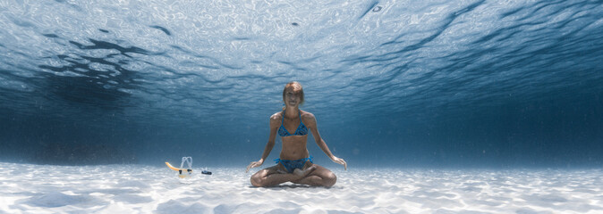 Slim yogi lady sits on the sandy sea bottom in the relaxed Lotus pose, smiles and looks at the...