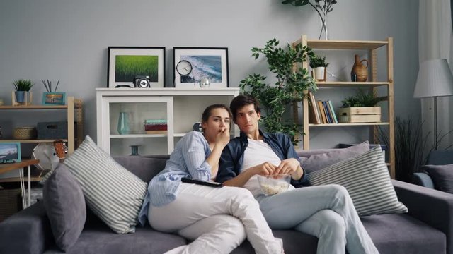 Zoom in of loving couple girl and guy watching horror film on TV eating popcorn closing eyes afraid of terrible movie. Youth and entertainment concept.