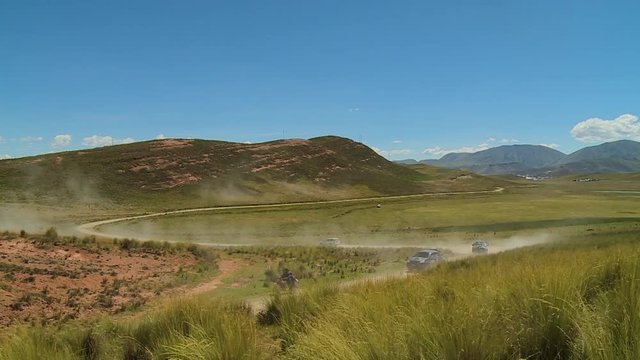 Wide still shot of a hilly landscape, s-curved country road with moving vehicles, shrubs on remote hills, and green grass cover on the lower plains, Peru.