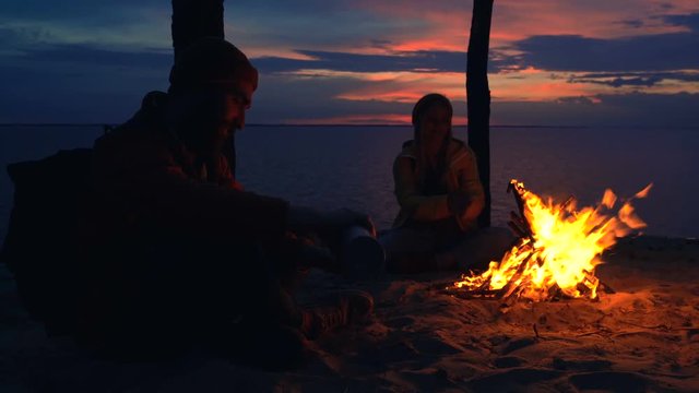 Couple of travelers sitting near the camfire. Young man pouring tea from thermos and giving it to his girlfriend.