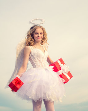 Portrait of a Cupid angel with gift. Girl angel with halo in white angel dress. Angel girl against sunny sky. Enjoying magic moment. Little goddess with white wings alone on blue sky background.