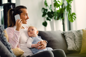 Young mother communicating on cell phone while enjoying with baby boy at home.