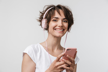 Young pretty excited happy woman posing isolated over white wall background using mobile phone listening music with earphones.