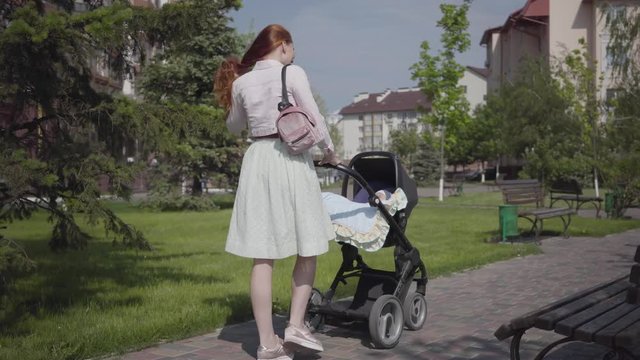 Beautiful red-haired woman walking on the walking along a stone walkway with a pram in the park. The lady enjoying the sunny day with her baby outdoors. Young mother walking with a child