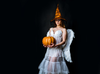 Angel Woman posing with Pumpkin. Young girl in witch Halloween hat on party over black background....
