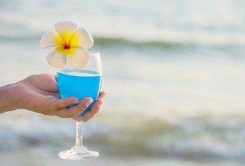Closeup woman hand holding cocktail glass decoration with plumeria flower with beach background - happy relax celebration vacation in sea nature concept