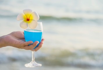 Fototapeta na wymiar Closeup woman hand holding cocktail glass decoration with plumeria flower with beach background - happy relax celebration vacation in sea nature concept