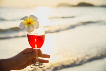 Closeup woman hand holding cocktail glass decoration with plumeria flower with beach background - happy relax celebration vacation in sea nature concept