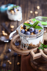 Healthy breakfast of blueberry parfaits made with fresh fruit, Greek yogurt, granola and mint leaves over a rustic cake stand.  Selective focus on glass jar in front with blurred background.