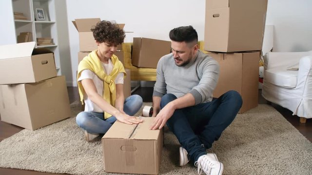 Full shot of middle-aged Caucasian man and woman sitting together on carpet at home and packing cardboard boxes with sticky tape gun to put things away for storage, while smiling and chatting