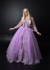 full length portrait of a blonde girl wearing a fantasy fairy inspired costume,  long purple ball gown with fairy wings,   standing pose on a dark studio background.