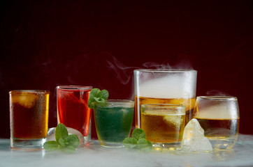 Different alcoholic cocktails with ice and mint with light smoke on a red background