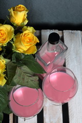 A large bouquet of yellow roses in a glass vase. Nearby is a wooden box. On the box is a bottle of liquor and glasses with a drink.
