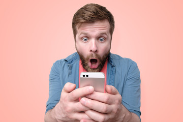 A surprised, shocked bearded man holds a smartphone in his hands and looks at the display with his mouth open and big eyes. Isolated on a coral background. Trendy colors.