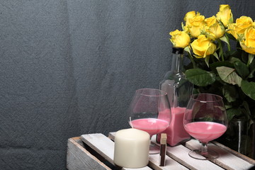 A large bouquet of yellow roses in a glass vase. Nearby is a wooden box. On the box is a bottle of liquor and glasses with a drink. Candle and matches.