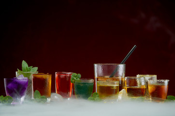 Color septet of alcoholic beverages in transparent glass glasses with ice and mint in smoke on a red background