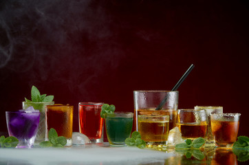 Color septet of alcoholic beverages in transparent glass glasses with ice and mint in smoke on a red background